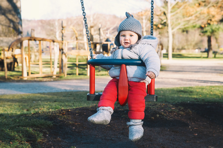 Portrait of cute baby dressed in warm clothes on swing at playground on sunny day outdoors.の写真素材