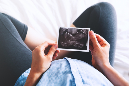 Pregnant woman keeping hand on belly and holding ultrasound image at home interiors. Pregnancy, parenthood, preparation and expectation concept. Close-up, copy space, indoors.の写真素材