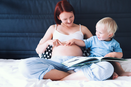 Pregnant mother and son are talking and reading book together in bed at home. Little child boy looking at her mother pregnant tummy. Pregnancy, family, parenthood, preparation and expectation concepts.の写真素材