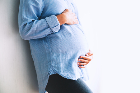 Pregnant woman holds hands on belly at home interiors. Pregnancy, parenthood, preparation and expectation concept. Close-up, copy space, indoors.の写真素材