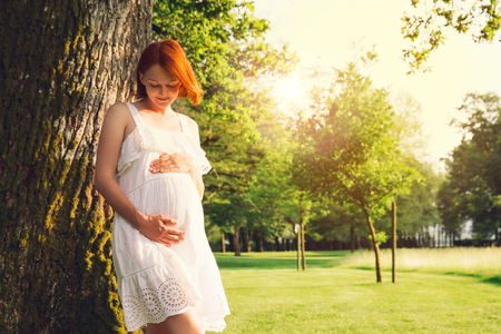 Beautiful pregnant woman in white dress on nature, outdoors. Expectant mother holds hands on belly on natural background of green grass in park at summer. Pregnancy, expectation, new life concepts.の写真素材