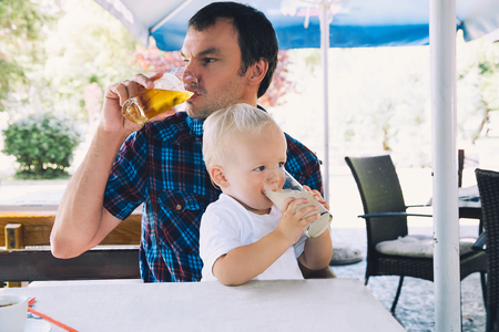 Father drinks beer, child drinks juice in a beach bar. Dad and son are best friends! Family spend time together in a cafe, restaurant in a summer day. Lifestyles, Family, Vacation concept.の写真素材