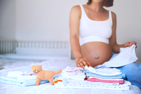 Pile of baby clothes, necessities and pregnant woman on bed in home interior of bedroom. Pregnant woman is getting ready for the maternity hospital, packing baby stuff. Pregnancy, birth concept.