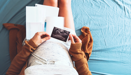 Pregnant woman makes notes, looking at ultrasound image and medical documents. Concept of pregnancy, health care, gynecology, medicine. Mother waiting of baby. Close-up, copy space, indoors.の写真素材