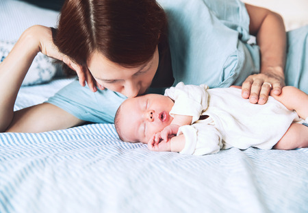 Mother kissing her newborn baby. Young beautiful mom lying at bed with a cute little sleeping child. New born baby's first days of life in family at home. Loving mother looks at asleep infant.の写真素材