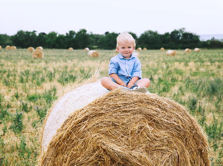 Cute toddler boy sitting on haystacks at wheat field. Happy child at summer day on nature, outdoors. Family, childhood background.の写真素材