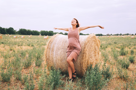 Beautiful pregnant woman in dress on nature raised arms on background of wheat field with haystacks at summer day.の写真素材