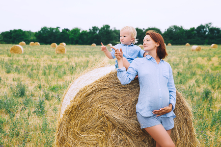Beautiful pregnant woman and her cute toddler son having fun on wheat field with haystacks at summer day on nature, outdoors.の写真素材