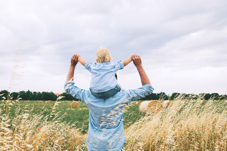 Happy father and son. Family concept. Man and his little cute kid boy having fun on wheat field with haystacks at summer day, outdoors. Dad carrying child on his back with raised arms up on nature.の写真素材