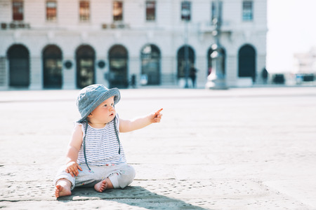 Baby girl sitting on a european city street and pointed finger up.  Little child outdoors in a town. Architecture of Trieste with pedestrian, people, Italy, Europe.の写真素材