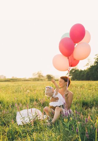 Baby girl in wicker basket with balloons in sunlight at sunset. Mother and child outdoors. First birthday. Family celebrates one year old baby on nature. Photo of healthy, dreams, vacation, holidaysの写真素材