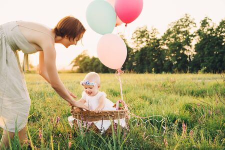 Baby girl in wicker basket with balloons in sunlight at sunset. Mother and child outdoors. First birthday. Family celebrates one year old baby on nature. Photo of healthy, dreams, vacation, holidaysの写真素材