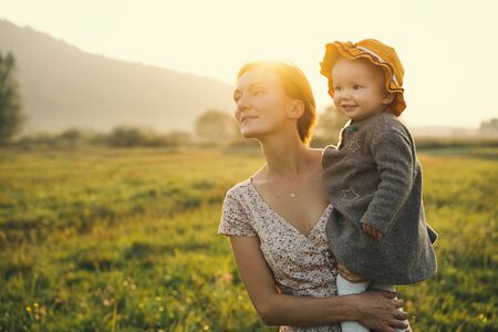 Young mother and baby girl in sunlight at sunset on nature outdoors. Mother and child in meadow in early autumn. Photo of natural parenting, dreams, family values, sustainable lifestyle.の写真素材