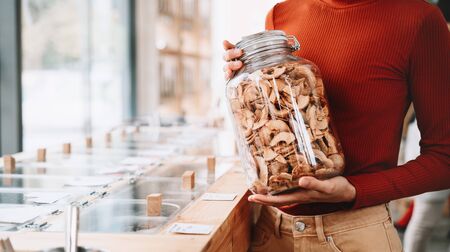 Concept of zero waste shop. Woman holding large glass jar with groceries on interior background of plastic free grocery store. Minimalist vegan style girl buying foods without plastic packaging.の写真素材