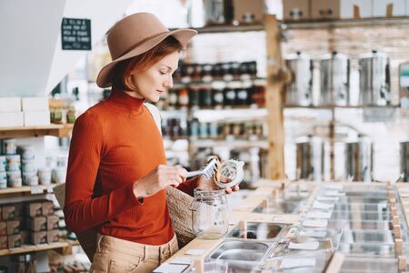 Young woman doing shopping with wicker bag and glass jars  in plastic free grocery store. Girl is buying products in zero waste shop. Concept of sustainable small businesses or minimalist lifestyle.の写真素材