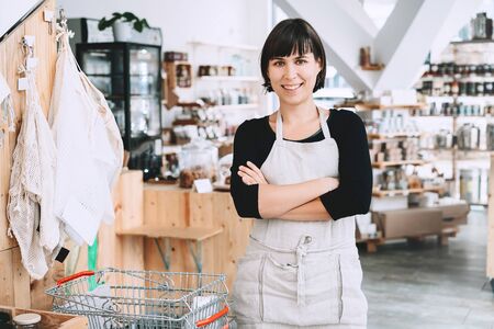 Portrait of owner of sustainable small local business. Female seller assistant of zero waste shop on interior background of shop. Smiling young woman in apron standing in plastic free grocery store.の写真素材