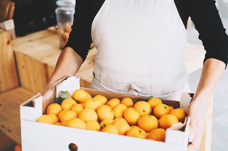 Close-up of hands holding cardboard box of organic tangerines in zero waste shop. Seller assistant with goods in plastic free store.の写真素材