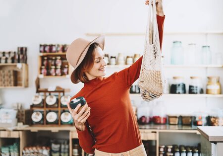 Minimalist vegan style young woman with cotton reusable net mesh bag and reusable glass coffee cup on background of goods of zero waste shop. Shopping without plastic packaging in plastic free store.の写真素材