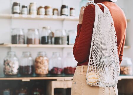 Minimalist vegan style young woman with cotton reusable net mesh bag on background of goods of zero waste shop. Shopping without plastic packaging in plastic free grocery store.の写真素材