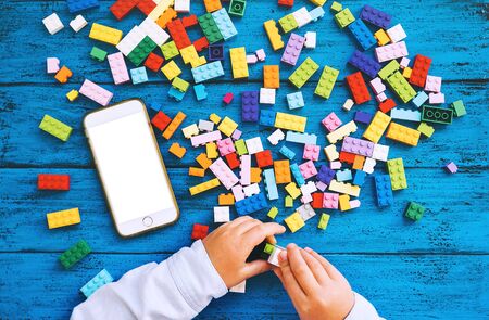 Background with child playing hands with colorful toy bricks or plastic blocks and mobile phone with blank white desktop screen. Kids leisure and education at home or class, learning and development.の写真素材