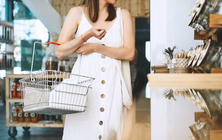 Shopping at local small business. Woman with grocery products in reusable glass jars inside metal basket in zero waste shop. Girl in white linen dress buying goods in sustainable plastic free store.の写真素材