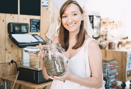 Girl holds glass jar with hemp seeds. Woman with cotton reusable bag chooses and buys products in zero waste shop. Weighing dry goods in plastic free grocery store. Eco shopping at local businessの写真素材