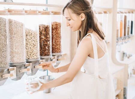 Woman with cotton bag and glass jar buying in zero waste shop. Dispensers for cereals, grains, nuts in plastic free grocery store. Sustainable shopping at small local businesses. Eco Bio Organic Foodの写真素材