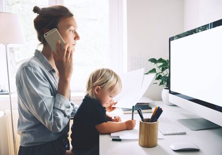 Modern mom balances between work and child home schooling on sick leave or quarantine. Mother works from home with kid. Woman responding on phone calls, working on computer with a toddler on her lap.の写真素材