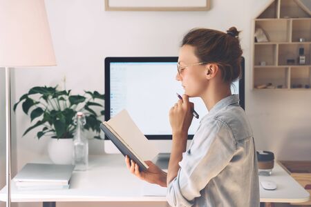 Woman freelancer indoors home office concept. Girl wearing in glasses works with new startup project. Female thinking and writing in planner or diary at workplace of minimalist style with computer.の写真素材