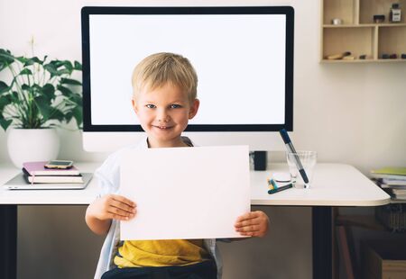 Preschool child boy holding in hands blank white paper on background of computer with blank white screen in home interior. Children online learning, distance education, development. Kids homeschoolingの写真素材