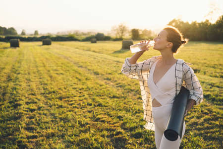 Image of yoga inspiration on nature. Young woman with yoga mat gets ready to practicing yoga outdoor. Beautiful female with slim, strength body in white clothes. Healthy and conscious lifestyles.の写真素材