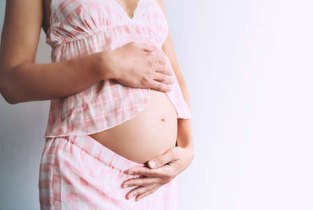 Young pregnant woman in home pajamas holds hands on belly on white background. Mother waiting for baby birth. Women prepare for maternity. Concept of pregnancy, prenatal period, maternal health.の写真素材