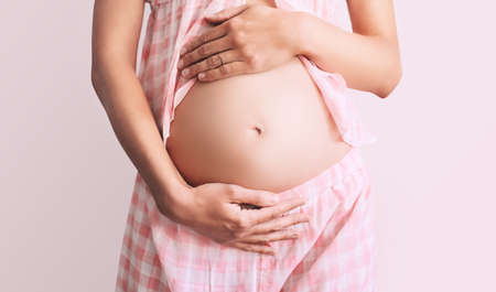 Young pregnant woman in home pajamas holds hands on belly on white background. Mother waiting for baby birth. Women prepare for maternity. Concept of pregnancy, prenatal period, maternal health.の写真素材