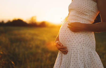 Young pregnant woman in white dress holds hands on her belly on green nature background. Close-up of...