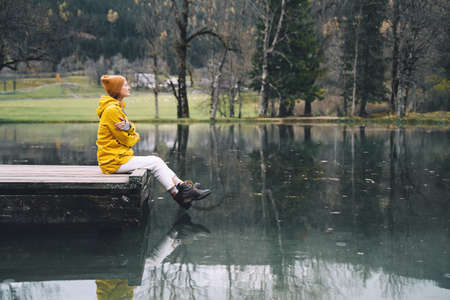 Girl in yellow raincoat looking at mountain lake in Jezersko. Adventure travel in Slovenia. Autumn or Winter in Europe. Woman sits on wooden bridge on nature background with Alps. Freedom Lifestyle.の写真素材