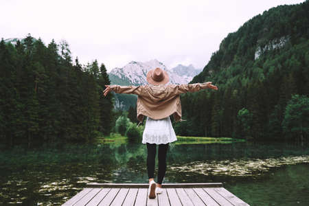 Young girl enjoying beauty of nature looking at mountain lake in Jezersko. Adventure travel in Slovenia, Europe. Woman stands with raised arms on wooden bridge on background with forest and Alps.の写真素材