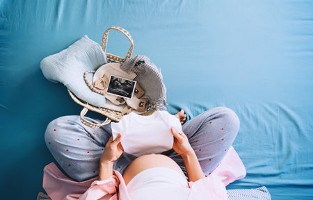 Pregnant woman holding baby bodysuit preparing to child birth during pregnancy. Mother with wicker basket with stuff for baby and ultrasound image. Concept local eco children products, baby shower.の素材 [FY310162439991]