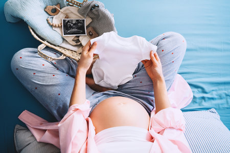 Pregnant woman holding baby bodysuit preparing to child birth during pregnancy. Mother with wicker...