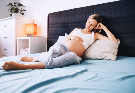 Beautiful pregnant woman holds hands on her belly and lying in bed at home, indoors. Young loving mother waiting of a baby. Concept of pregnancy, maternity, healthcare, gynecology.の写真素材