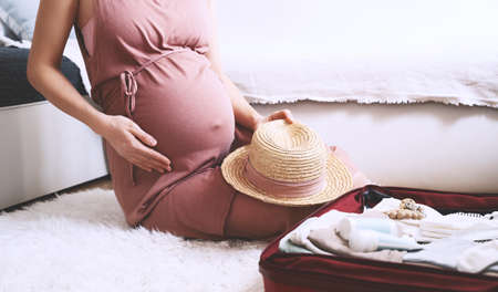 Close-up belly of pregnant woman in dress with travel bag of clothes and necessities. Ð¡areful mother during pregnancy preparing and packing suitcase for maternity hospital getting ready for baby birth.の写真素材