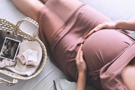 Pregnant woman in dress holds hands on belly. Mother with wicker basket with stuff for newborn, ultrasound image and teddy toy. Expectant mother waiting and preparing for baby birth during pregnancy.の写真素材