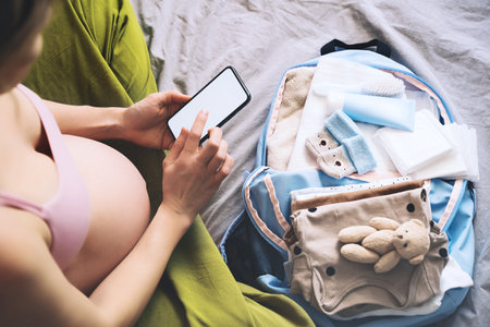 Pregnant woman getting ready for labor packing stuff for hospital, making notes or checklist in smartphone. Baby clothes, necessities for mother and newborn in maternity bag. Pregnancy concept.の写真素材