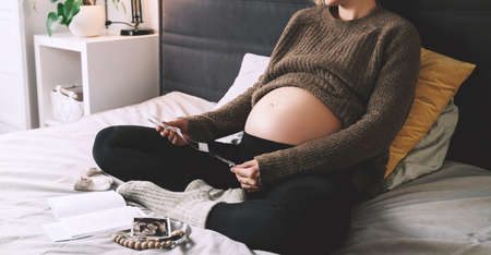Pregnant woman holding ultrasound image looking at medical test reports in cozy knitted sweater in bed at cold winter days. Expectant mother with pregnant belly waiting for baby. Concept of pregnancy.の写真素材