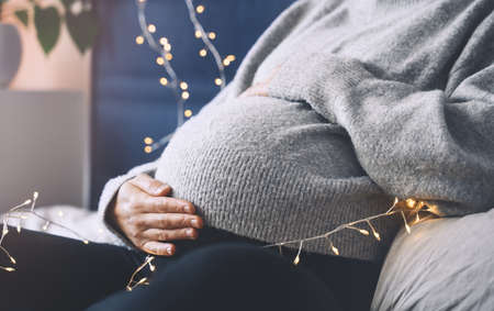 Close-up beautiful belly of pregnant woman in knitted sweater in light of garlands. Loving mother waiting a baby. Concept of pregnancy, maternity, baby shower party, holiday winter mood.の写真素材