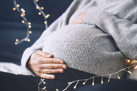 Close-up beautiful belly of pregnant woman in knitted sweater in light of garlands. Loving mother waiting a baby. Concept of pregnancy, maternity, baby shower party, holiday winter mood.の写真素材