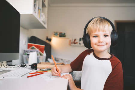 Child 7 years old is homeschooling or distance online education. Elementary school boy studying, does homework or creating at desk. Smiling kid in headphones near computer drawing or writing at home.の写真素材