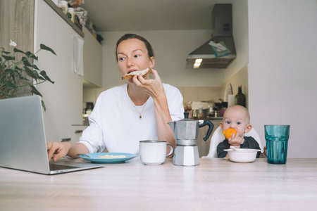Young woman studying or working online at home while having breakfast with her baby in kitchen. Millennial mother on maternity leave with child. Freelancer busy mom with laptop searching information.の写真素材