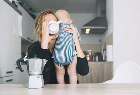 Modern young tired mom and little child after sleepless night. Exhausted woman with baby is sitting with coffee in kitchen. Life of working mother with baby. Postpartum depression on maternity leave.の写真素材