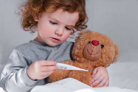 a little girl in pajamas sits on a bed with a brown teddy bear and holds a thermometer in her hand and looks at it, the concept of medicine.の写真素材