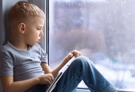a little fair-haired boy in a gray T-shirt and jeans sits on the windowsill, holds a tablet in his hands and looks sadly out the window, it's raining. social concept. stay homeの写真素材